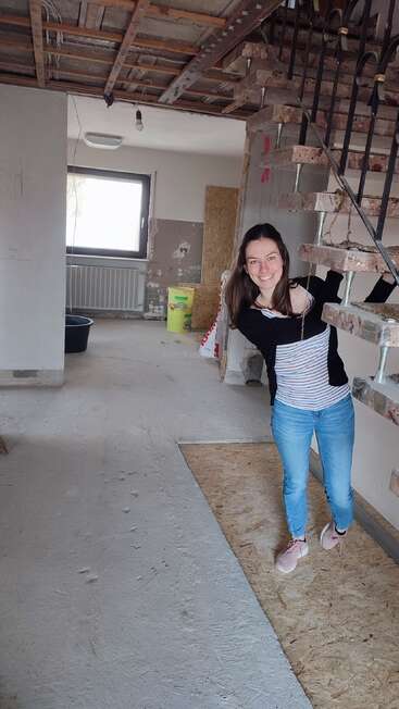 Una mujer sonriente posa con alegría en unas escaleras sin terminar de una casa en obras. Al fondo se ven el techo sin revestir, las paredes desnudas y los materiales de construcción.