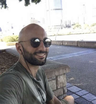 Um homem careca e barbudo, usando óculos escuros e fones de ouvido, sorri enquanto está sentado ao ar livre em um banco. Você pode ver prédios e árvores da cidade.