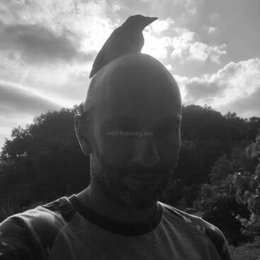 Eine Schwarz-Weiß-Fotografie eines Mannes im Freien, auf dessen kahlem Kopf ein Vogel thront. Der Himmel und die Bäume bilden einen dramatischen, natürlichen Hintergrund.