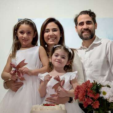 A happy family of four stands together, smiling. Two young girls in white dresses hold ribbons. Flowers and a cake are in the foreground, creating a festive atmosphere.