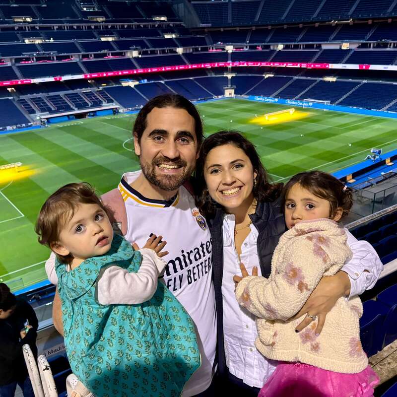 L'image montre une famille de quatre personnes posant pour une photo devant un terrain de football à l'intérieur d'un stade, le père portant un maillot de football.