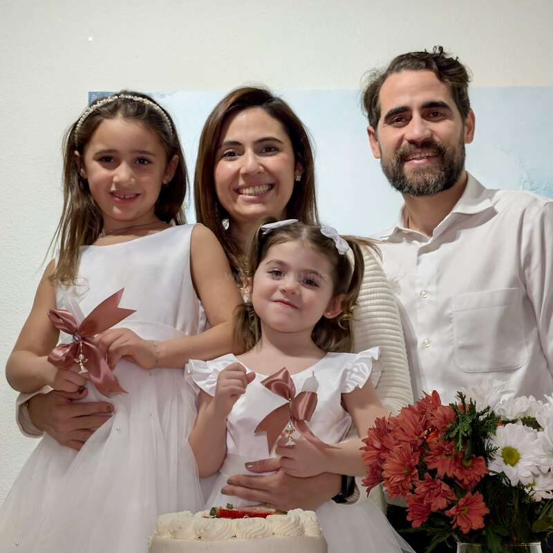 Eine glückliche vierköpfige Familie steht zusammen und lächelt. Zwei junge Mädchen in weißen Kleidern halten Bänder. Im Vordergrund stehen Blumen und ein Kuchen, die eine festliche Atmosphäre schaffen.