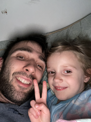 Das Bild zeigt einen Mann und ein junges Mädchen, die im Bett liegen und in die Kamera lächeln, während das Mädchen mit ihren Fingern ein Friedenszeichen macht.