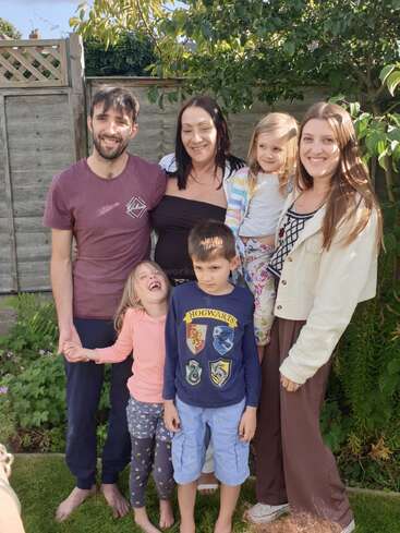 Das Bild zeigt eine sechsköpfige Familie, die für ein Foto in einem Hinterhof posiert, mit zwei Erwachsenen und vier Kindern, umgeben von üppigem Grün und einem Holzzaun.