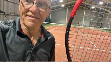 Um homem usando óculos sorri para uma selfie em uma quadra de tênis coberta, segurando uma raquete de tênis em primeiro plano. Outro jogador pode ser visto em segundo plano.