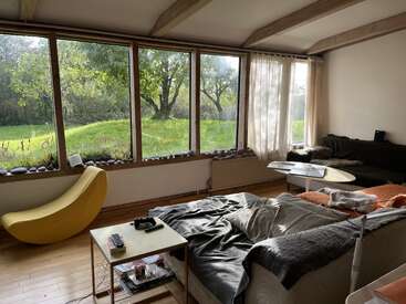 Essa sala de estar aconchegante tem janelas grandes com vista para um jardim verdejante, sofás confortáveis, uma cadeira amarela exclusiva e uma mesa de centro com livros e controles remotos.