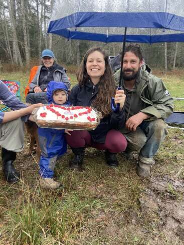 Una familia celebra al aire libre un día lluvioso, con un paraguas y una tarta de cumpleaños decorada con frambuesas. Todos sonríen, a pesar del entorno embarrado y húmedo.