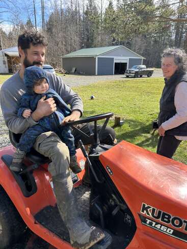 Un hombre que sostiene a un niño en brazos está sentado en un tractor Kubota y sonríe a una mujer mayor que se encuentra de pie cerca de él. Es un día soleado en un entorno rural.