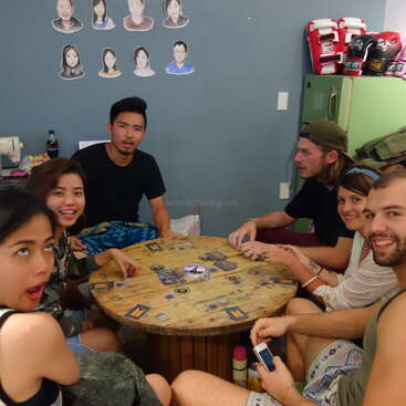 La imagen muestra a un grupo de personas jugando a un juego de mesa, con una mesa y sillas al fondo, y algunas personas sentadas alrededor de la mesa. El ambiente parece relajado y social.