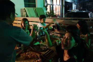 Un grupo de amigos disfruta de una reunión nocturna al aire libre, compartiendo pizza, bebidas y risas, brindando y celebrando juntos bajo una iluminación verde.