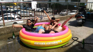 Tres personas se relajan y posan con entusiasmo en una pequeña piscina hinchable en un día soleado, rodeadas de coches aparcados, patinetes y botellas de vidrio vacías en el exterior.