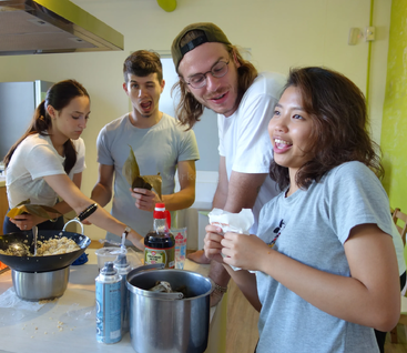 Cuatro jóvenes cocinan juntos en una cocina iluminada, riendo y divirtiéndose. Hay varios ingredientes y utensilios esparcidos por la encimera.