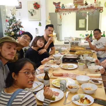 Un grupo de amigos disfruta de una comida festiva alrededor de una mesa, rodeados de adornos navideños, pizza, bebidas y risas en un acogedor ambiente casero.