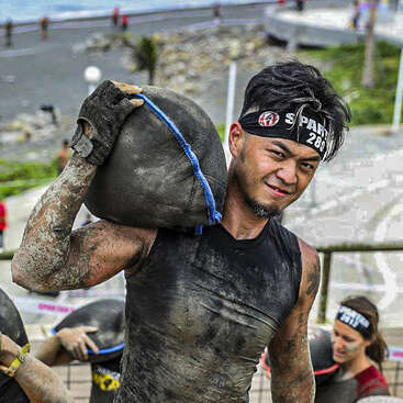 Un athlète boueux participe à une course d'obstacles en plein air, portant un lourd sac de sable sur l'épaule, muni de gants et d'un bandeau Spartan, l'air déterminé et concentré.