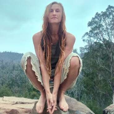 Uma mulher com longos dreadlocks agacha-se descalça em uma rocha, cercada por árvores e montanhas. Sua roupa é casual e ela parece serena ao ar livre.