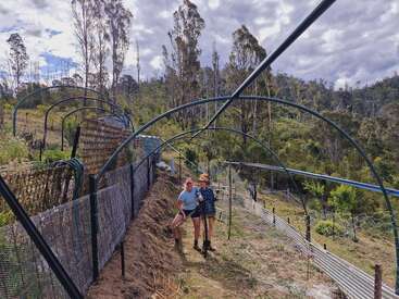 Deux femmes se tiennent sous des arches métalliques dans un jardin, entouré de clôtures et d'arbres. Le ciel est partiellement nuageux et le paysage est vallonné et verdoyant.