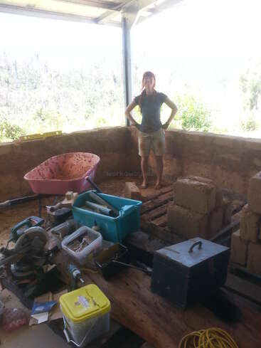 Une femme se tient à l'intérieur d'une structure partiellement construite avec des murs en terre, entourée d'outils de construction, de matériaux, d'une brouette et de blocs, sous un toit en métal.