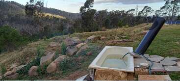 Une baignoire rustique d'extérieur est posée sur une colline herbeuse, entourée de rochers. Un chauffe-eau solaire y est fixé. Des montagnes boisées et des arbres créent une toile de fond pittoresque.