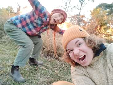 Deux personnes vêtues de vêtements chauds, de bonnets et de bottes posent avec enthousiasme pour un selfie en plein air, avec un grand feu lumineux brûlant à l'arrière-plan au milieu de l'herbe sèche.