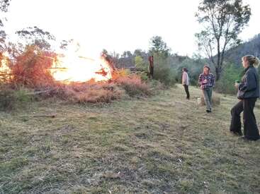 Trois personnes se tiennent sur un terrain herbeux et observent un grand feu de joie qui brûle intensément. La zone est entourée d'arbres et de buissons, sous un ciel ouvert au crépuscule.