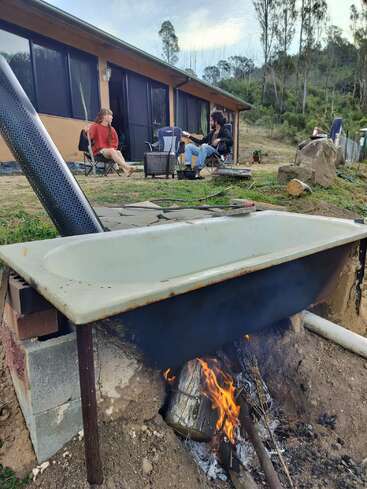 Deux personnes sont assises et discutent à l'extérieur d'une maison, tandis qu'une grande baignoire en métal est chauffée sur un feu ouvert à proximité, entourée d'arbres et d'un environnement terreux.