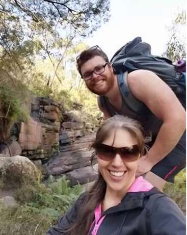 Um homem e uma mulher sorriem ao ar livre, usando óculos escuros e roupas casuais, aproveitando uma caminhada numa área rochosa e arborizada, com vegetação exuberante e a luz do sol a penetrar entre as árvores.