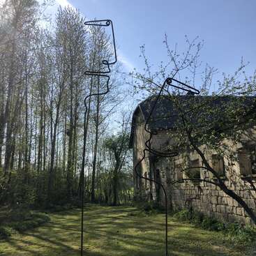Das Bild zeigt eine ruhige Szene mit einem Steingebäude mit dunklem Dach, umgeben von Bäumen und einer Grasfläche, auf der zwei schwarze Metallskulpturen im Vordergrund zu sehen sind.