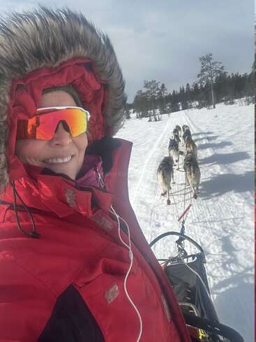 Una persona con abrigo rojo de invierno y gafas de sol reflectantes sonríe mientras conduce un trineo tirado por perros por un sendero nevado con huskies, rodeado de árboles.