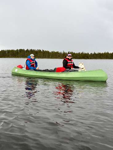 Deux personnes portant des gilets de sauvetage et un chien blanc sont assis dans un canoë vert sur une eau calme, avec un rivage boisé sous un ciel gris nuageux.