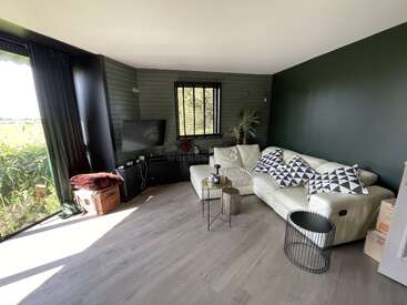 La imagen muestra un acogedor salón con un sofá blanco, televisor y suelo de madera, con una ventana con vistas al exterior.