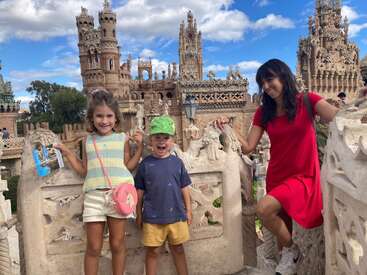Une femme et deux enfants posent joyeusement devant une architecture en pierre complexe avec de hautes tours, sous un ciel bleu vif parsemé de nuages cotonneux.