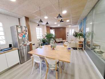 Esta imagem mostra uma cozinha moderna e aberta, integrada à sala de estar, com mesas de madeira, sofás aconchegantes, plantas de interior, luminárias suspensas, janelas amplas e um ambiente claro e acolhedor.