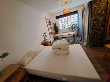 Um quarto aconchegante e moderno com uma cama branca, móveis de madeira, uma mesa, plantas verdes, luz natural suave e cortinas estampadas, criando um ambiente confortável e acolhedor.