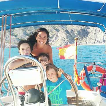 The image shows a woman and three children on a boat in the water, with a rocky coastline in the background and a life ring visible.