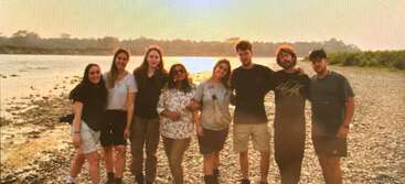 La imagen muestra a un grupo de personas de pie, con una mezcla de hombres y mujeres, todos mirando a la cámara, con el telón de fondo de una masa de agua y un cielo despejado.