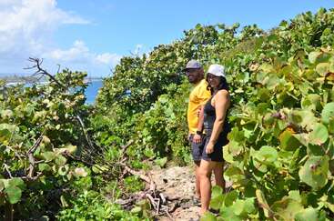 L'image représente un homme et une femme debout sur un chemin rocailleux, entourés d'une végétation luxuriante, avec un plan d'eau serein et un ciel bleu en arrière-plan.