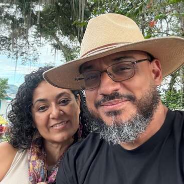 A smiling couple poses outdoors. The woman has curly hair and wears a colorful scarf, while the man wears glasses, a beige hat, and a black shirt.