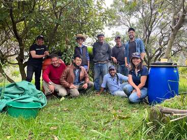 Eine Gruppe von neun Leuten steht draußen, umgeben von Bäumen und viel Grün. Sie lächeln glücklich, einige knien, andere stehen, und neben ihnen stehen Fässer und Behälter, was auf Teamarbeit hindeutet.