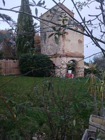 An old, rustic stone tower stands in a grassy garden, surrounded by trees and shrubs. Branches frame the scene, evoking a peaceful, rural atmosphere.