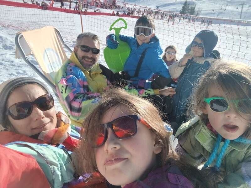 A family enjoys a sunny day in the snow, wearing colorful winter gear and sunglasses, smiling, relaxing, and having fun together at a ski resort.