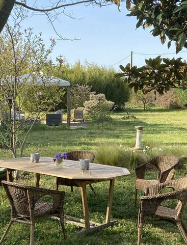 Uma cena encantadora no jardim com uma mesa de madeira, quatro cadeiras de vime e flores frescas. Cercada por árvores, vegetação e um tranquilo espaço ao ar livre sob um céu azul.