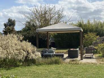 Um aconchegante gazebo ao ar livre fica rodeado por uma vegetação exuberante e arbustos floridos, com espreguiçadeiras e um sofá sob um toldo branco, sob um céu parcialmente nublado.