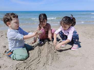 Três crianças estão brincando juntas em uma praia de areia, construindo um castelo de areia. As ondas do mar e o céu azul criam uma atmosfera pacífica e alegre ao fundo.