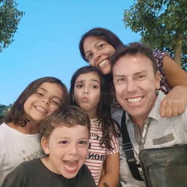 Uma família feliz de cinco pessoas posa ao ar livre, sorrindo brilhantemente para a câmera. As crianças fazem caretas brincalhonas, enquanto os pais ficam atrás, curtindo o momento de alegria juntos.