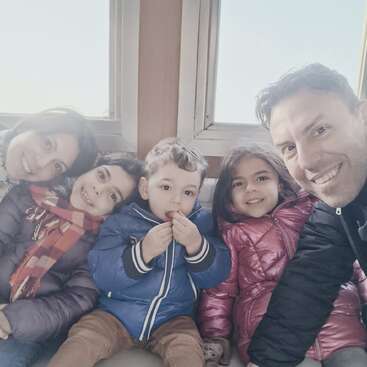 Uma família feliz de cinco pessoas senta-se junta, sorrindo calorosamente. Os pais e os três filhos usam casacos de inverno aconchegantes, aproveitando um dia claro e a companhia uns dos outros.