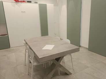 Esta imagem mostra uma sala de jantar minimalista com uma mesa retangular cinza, quatro cadeiras brancas, piso de ladrilho, armários verdes e brancos e um documento sobre a mesa.