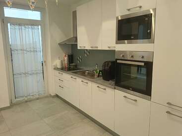 Esta moderna cocina cuenta con armarios blancos, horno empotrado, microondas, cafetera, fregadero de acero inoxidable, encimeras grises, suelo de baldosas y una puerta de cristal con cortinas.