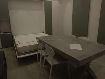 Una habitación moderna y minimalista con una cama abatible, una gran mesa de comedor gris, sillas blancas y una decoración sencilla. Sobre la mesa hay un único documento.