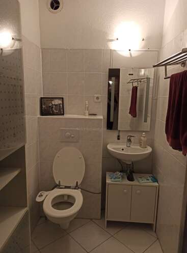 The image depicts a compact bathroom with white tiles, featuring a toilet, sink, and mirror, illuminated by two lights and a small cabinet with a towel rack.