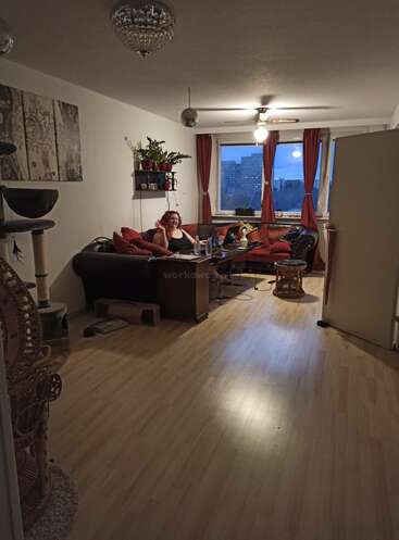The image depicts a woman sitting on a couch in a living room, surrounded by a cat tree, plants, and a window with red curtains, with a cityscape visible outside.